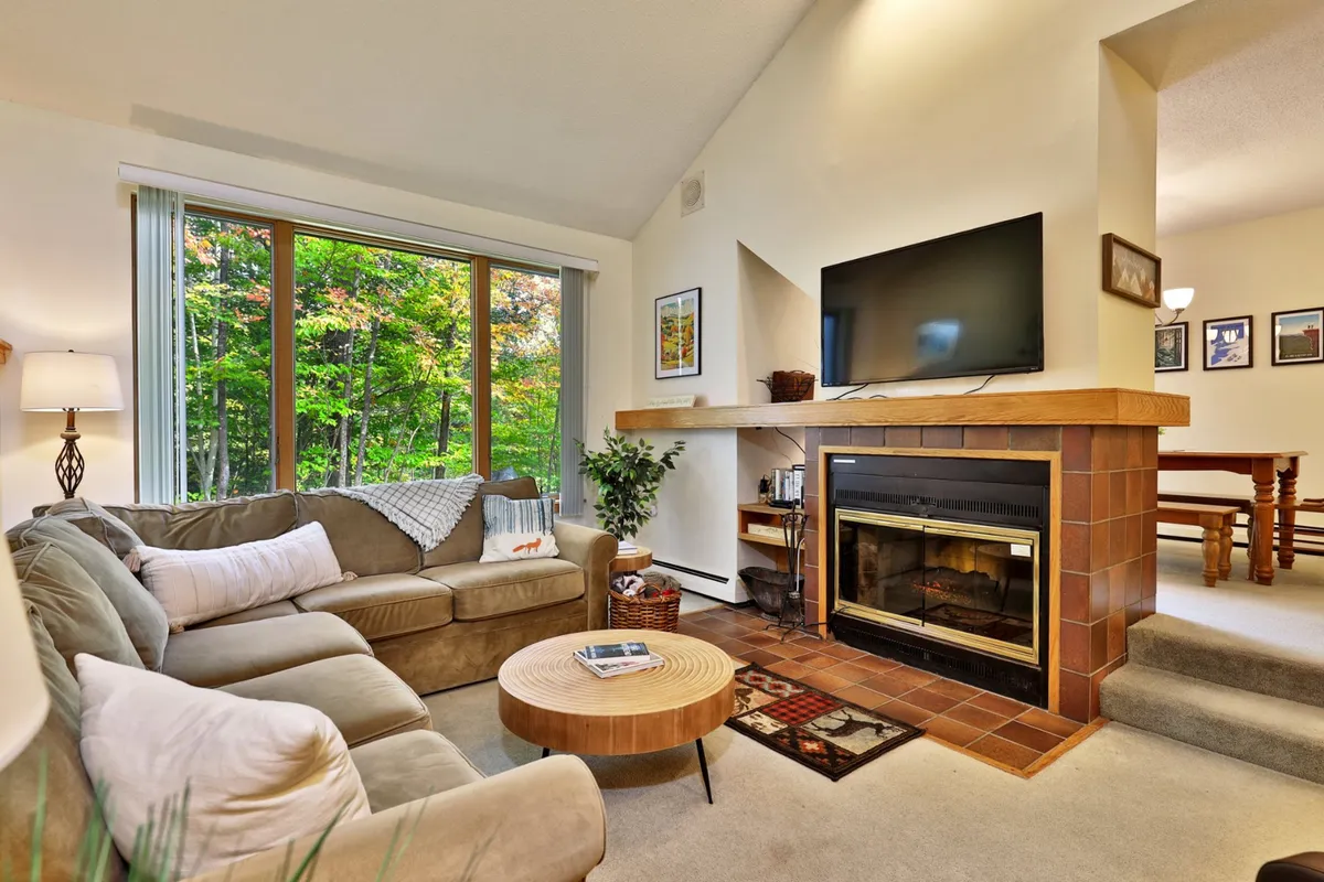 A warm, welcoming seating area featuring large windows that showcase tranquil nature views. A comfortable seating arrangement is oriented around a cozy fireplace and coffee table, creating an inviting space for relaxation or socializing. The natural light enhances the sense of peace and retreat.