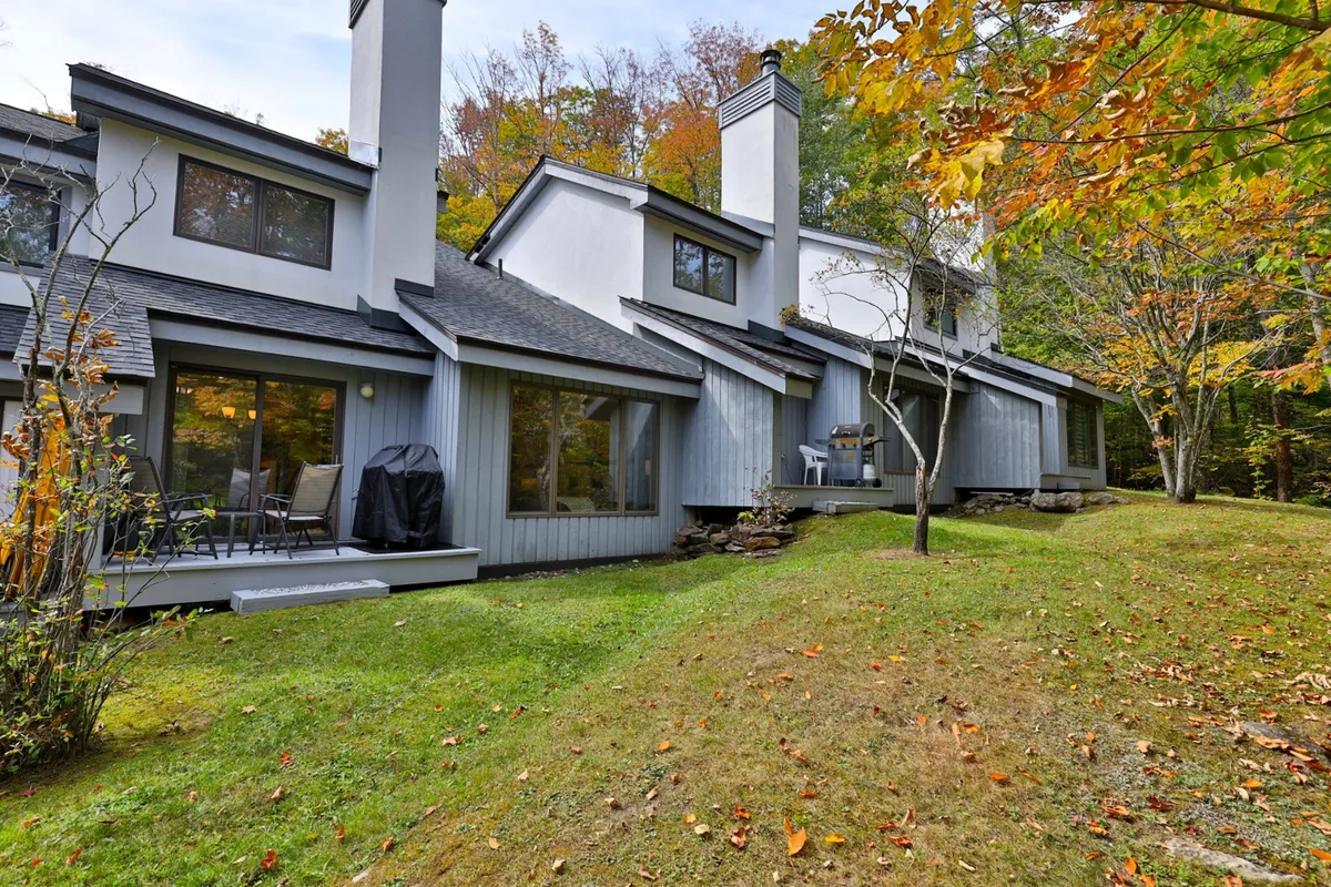 A two-story house with a spacious deck set amidst tall trees. The deck offers space for outdoor dining and gathering, while the surrounding foliage sets the scene for stargazing or seasonal views. The peaceful setting invites both social and solitary enjoyment.