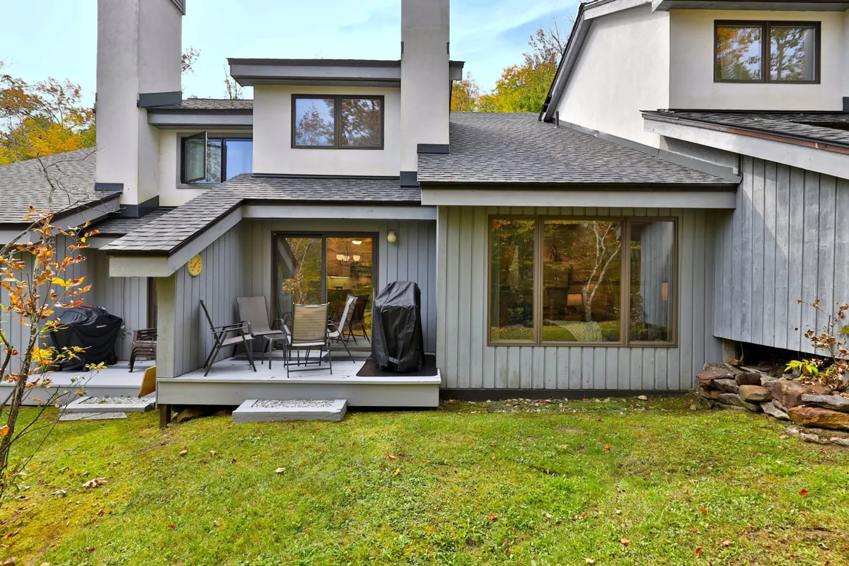 A private patio area furnished with comfortable outdoor seating, a BBQ grill, and surrounded by lush greenery. Large windows from the adjacent living room offer a seamless indoor-outdoor connection. The scene is set for outdoor dining, relaxing with friends, or soaking up the natural setting.