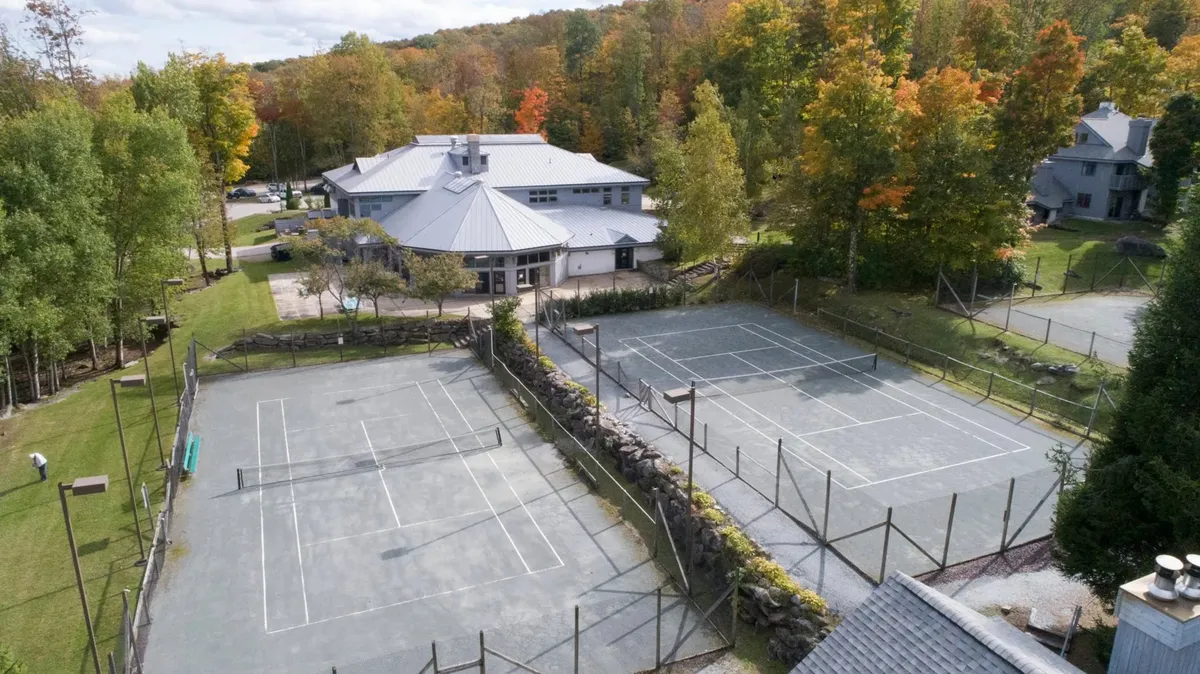 An inviting exterior view of the main building surrounded by mature trees. In the foreground, well-maintained tennis courts are visible, suggesting opportunities for outdoor activities and socializing. The scene is lively and offers a glimpse into the communal atmosphere outside the property.
