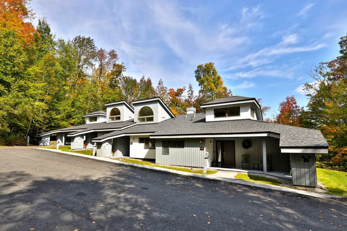 A modern condo exterior surrounded by colorful autumn foliage, a well-kept driveway, and hints of nearby natural trails. The building sits within a peaceful, wooded setting, suggesting immediate access to outdoor adventures.
