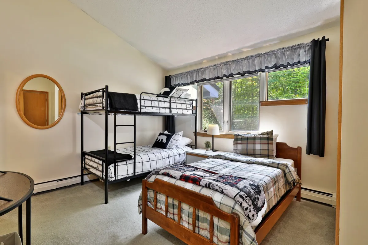 This image showcases a bedroom with a twin bed and a bunk bed, a large window for natural light, a round mirror, and a decorative lamp. The bunk bed is accessible by a ladder, which matches the section's mention of accommodations suitable for siblings or friends sharing space.