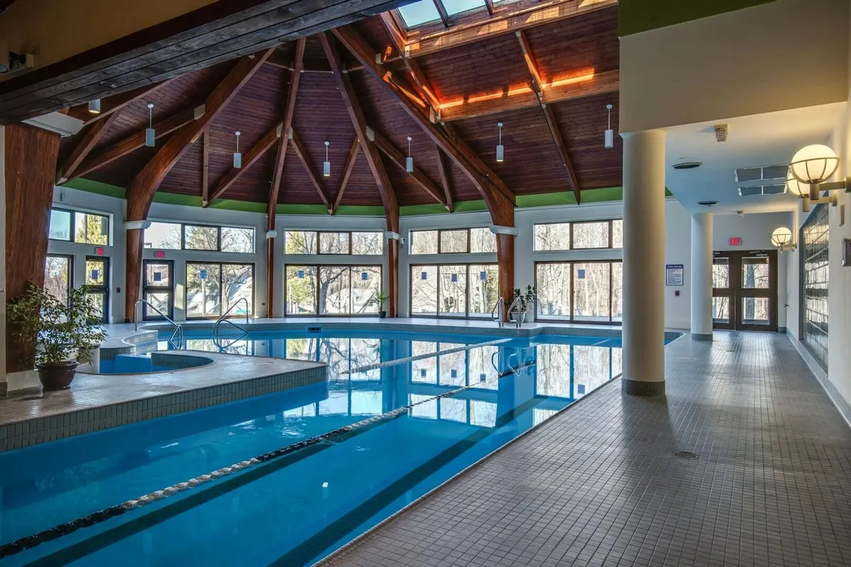 A bright and tranquil indoor swimming pool space illuminated by natural light streaming through large windows and overhead skylights. The wooden beams add warmth and architectural character, while the pool offers a serene spot for both relaxation and active swimming.